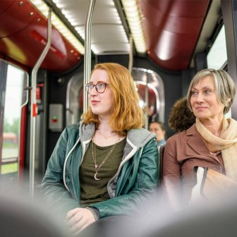 Eine junge Frau mit roten, mittellangen Haaren, einer schwarzen Brille und einer dunkelgrünen Lederjacke und eine ältere Frau mit kurzen, grauen Haaren, einem braunen Mantel und einem hellbraunen Schal sitzen in einer Stadtbahn der BSVG und blicken lächel