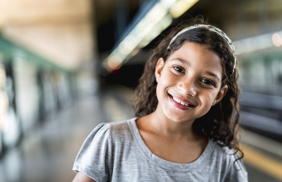 Ein Mädchen mit braunen, lockigen Haaren trägt ein graues T-Shirt, steht an einem Bahnsteig und lächelt fröhlich in die Kamera.