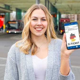 Eine blonde Frau in einer grauen Jacke steht vor einer Bushaltestelle und hält lächelnd ein Smartphone in die Kamera. Auf dem Smartphone ist das Deutschlandtticket-Logo zu sehen.