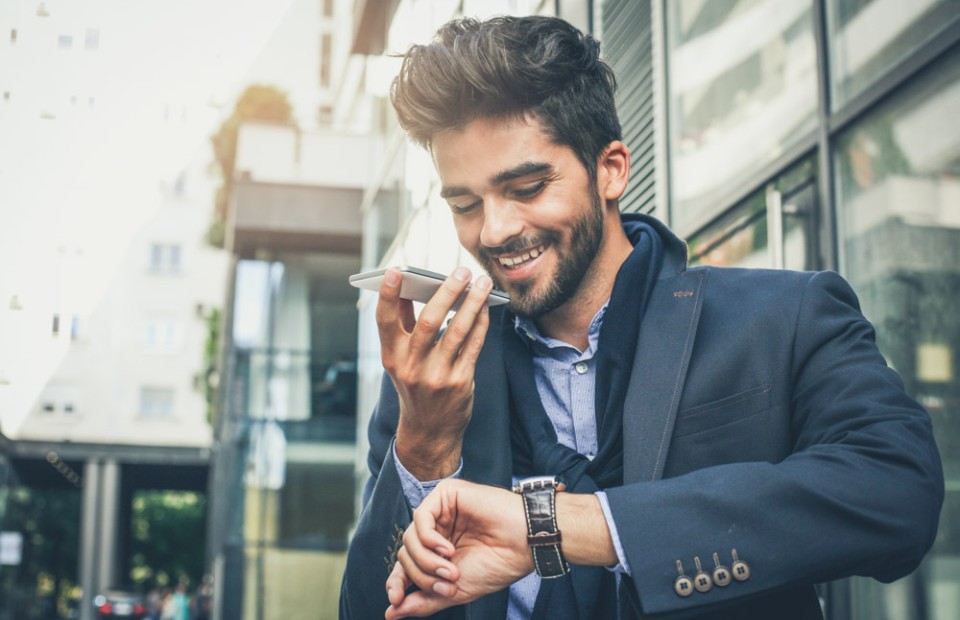 Ein Mann mit dunklen, kurzen Haaren und Bart trägt einen blauen Anzug, blickt auf seine Armbanduhr und spricht dabei freundlich lächelnd in sein Smartphone.