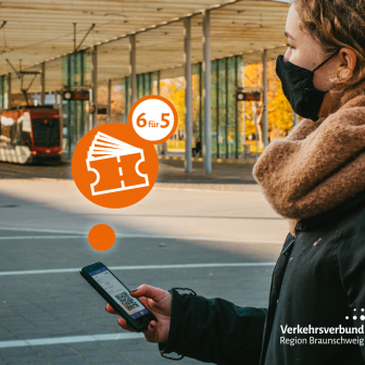 Eine Frau in Winterjacke und braunem Schal mit schwarzer Schutzmaske kauft am Braunschweiger Hauptbahnhof über die VRB-App ein Mehrfahrtenticket. Im Hintergrund hält eine Stadtbahn der BSVG an der Bahnhaltestelle.