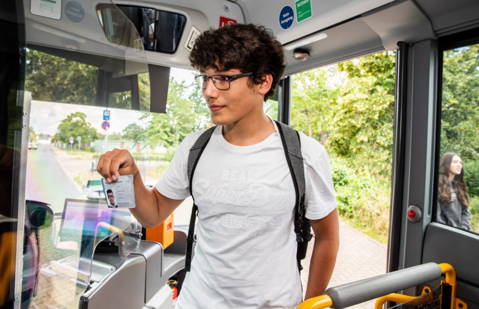 Ein Junge mit dunklen, kurzen Locken und schwarzer Brille trägt ein hellgraues T-Shirt und einen grauen Rucksack und zeigt beim Einsteigen in einen Bus seine Schüler-Jahreskarte vor.