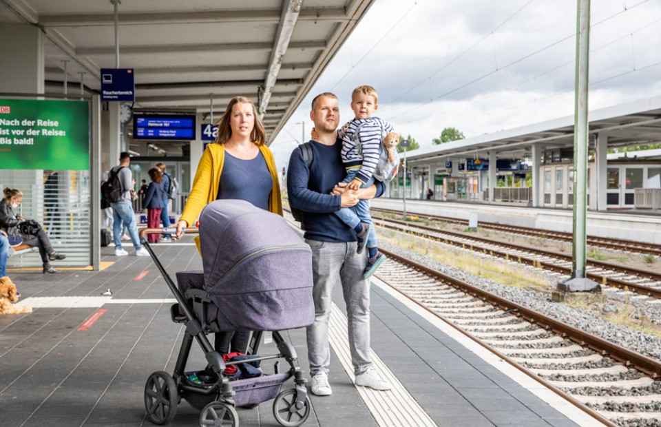 Eine Familie steht an einem Bahnsteig am Braunschweiger Hauptbahnhof. Der Mann trägt einen blauen Pullover, eine graue Jeans und einen Rucksack und hat seinen Sohn auf dem Arm. Die Frau trägt eine senfgelbe Jacke. Vor ihr steht ein grauer Kinderwagen, den