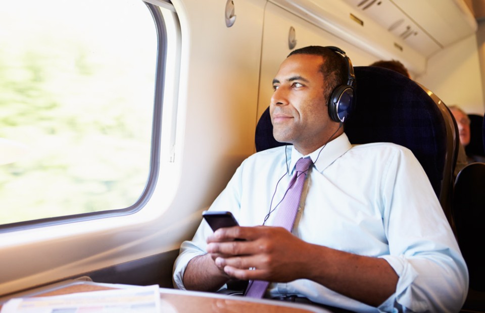 Ein dunkelhaariger Mann in einem weißen Hemd mit rosa Krawatte und hochgekrempelten Ärmeln sitzt im Zug und blickt aus dem Fenster. Er trägt Kopfhörer und hält ein Smartphone in den Händen.