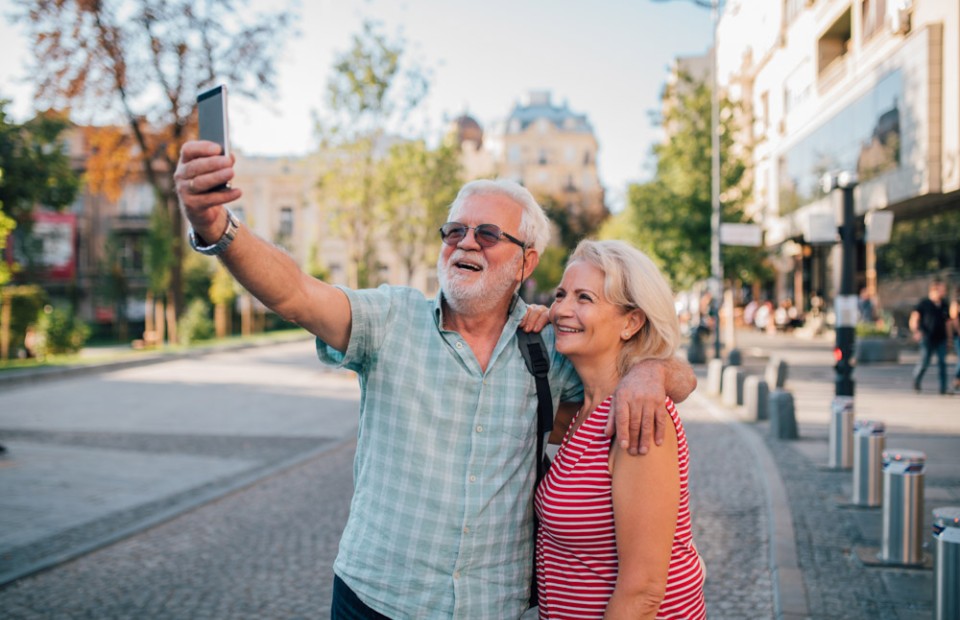 Ein älteres, glücklich lächelndes Paar geht an einem sonnigen Tag durch die Stadt und macht ein Selfie, auf dem es sich in den Armen hält.