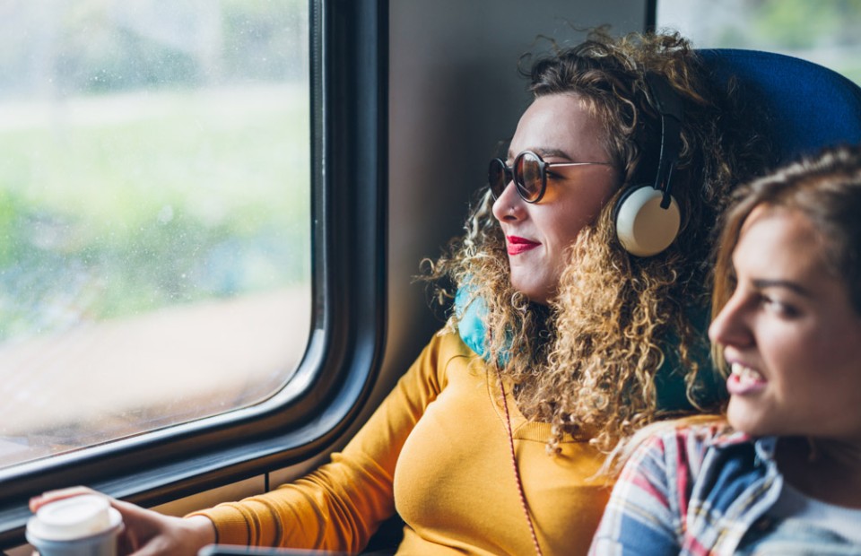 Zwei Frauen sitzen im Zug und blicken aus dem Fenster. Die links sitzende Frau trägt Kopfhörer und hält einen Kaffee in ihrer Hand.