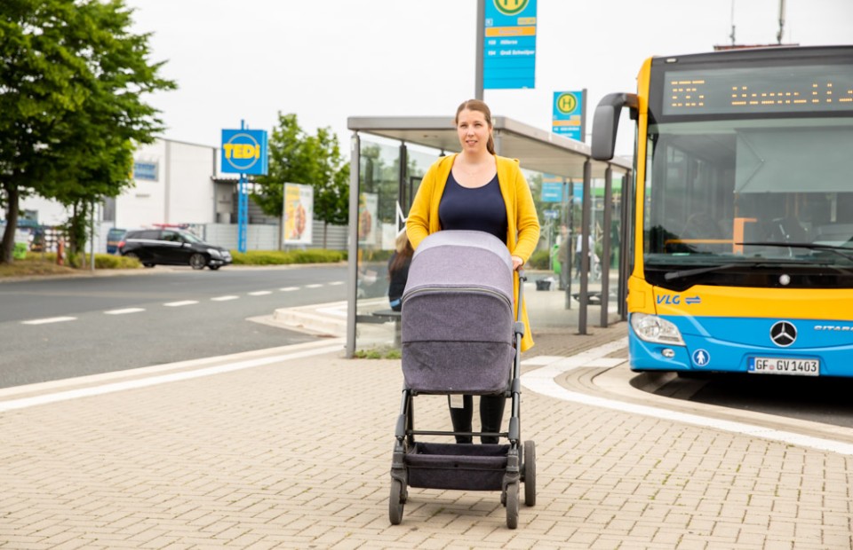 Eine dunkelhaarige Frau in senfgelber Jacke steht mit einem grauen Kinderwagen vor einer Haltestelle in Gifhorn. Hinter ihr steht ein blau-gelber Bus der VLG.