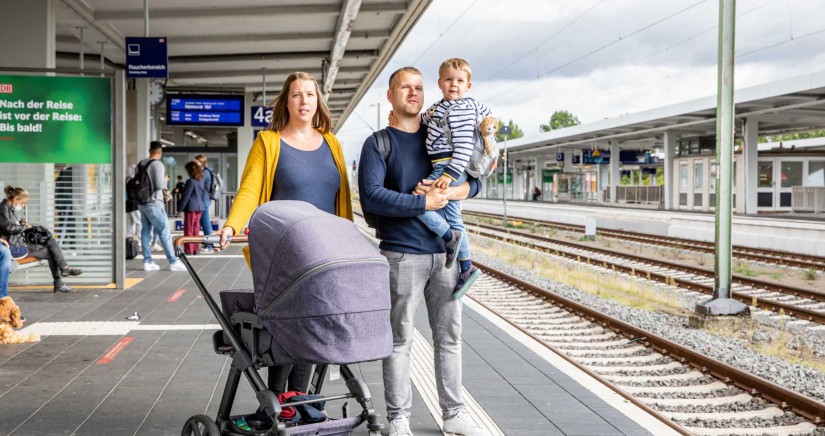 Eine Familie steht am Bahnhof und wartet auf den Zug. Die Frau trägt eine gelbe Jacke und schiebt einen Kinderwagen. Der Mann hat das Kind auf dem Arm.