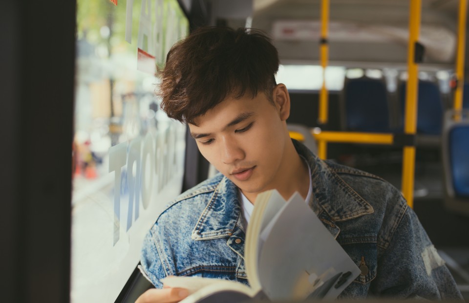 JEin junger Mann mit dunklen Haaren und Jeansjacke sitzt in einem Bus an einem Fensterplatz und liest ein Buch.