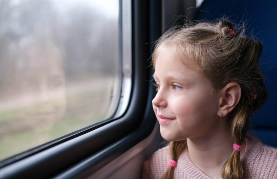 Ein blondes Mädchen mit zwei geflochtenen Zöpfen sitzt in einem Zug und schaut lächelnd aus dem Fenster.