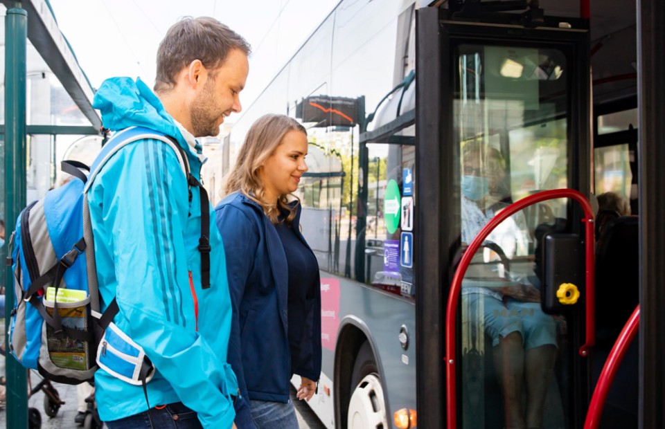 Eine blonde Frau in dunkler Softshell-Jacke und ein dunkelhaariger Mann mit Drei-Tage-Bart, der eine türkisfarbene Regenjacke und einen blauen Rucksack trägt, steigen lächelnd gemeinsam in einen Bus ein.