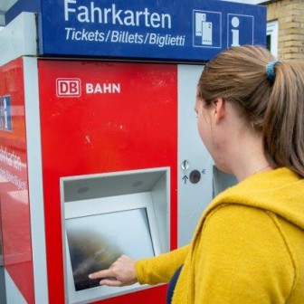 Eine Frau in einem gelben Pulli kauft an einem Automaten der Deutschen Bahn ein VRB-Ticket.
