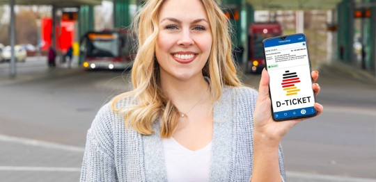 Eine blonde Frau in einer grauen Jacke steht vor einer Bushaltestelle und hält lächelnd ein Smartphone in die Kamera. Auf dem Smartphone ist das Deutschlandtticket-Logo zu sehen.