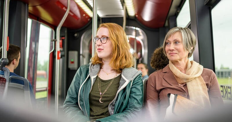 Eine junge Frau mit roten, mittellangen Haaren, einer schwarzen Brille und einer dunkelgrünen Lederjacke und eine ältere Frau mit kurzen, grauen Haaren, einem braunen Mantel und einem hellbraunen Schal sitzen in einer Stadtbahn der BSVG und blicken lächel