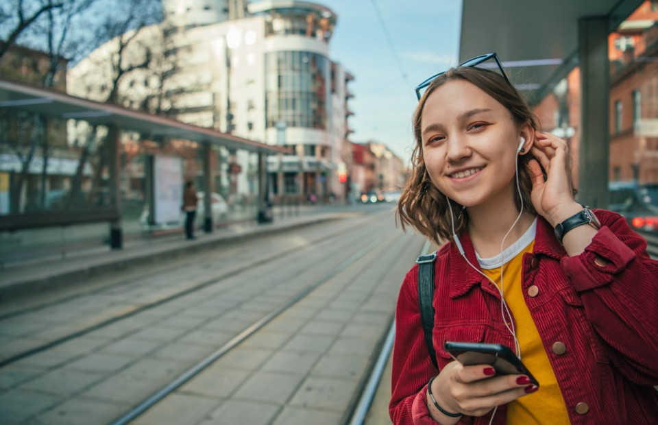 Ein Mädchen mit dunklen, mittellangen Haaren und roter Kord-Jacke wartet an einer Straßenbahnhaltestelle auf die Bahn. Sie trägt Kopfhörer und hält ihr Smartphone in der einen Hand, während sie sich mit der anderen Hand die Haare hinter ihr Ohr streicht.