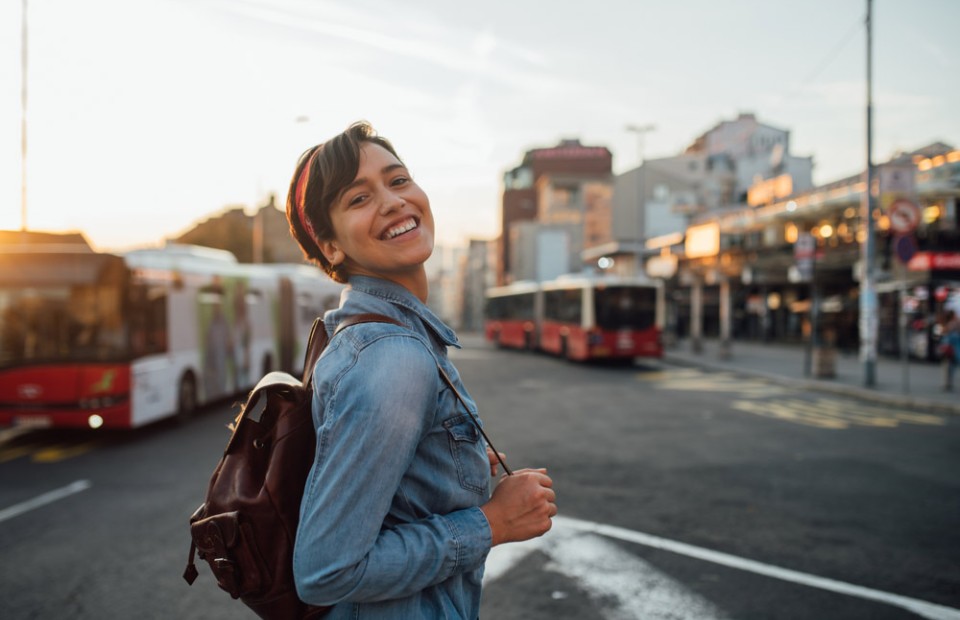 Eine Frau mit braunen, kurzen Haaren und Haarband geht zu einer Bushaltestelle und lacht in die Kamera. Sie trägt eine Jeansjacke und einen schwarzen Rucksack. Im Hintergrund sind zwei Busse zu sehen.