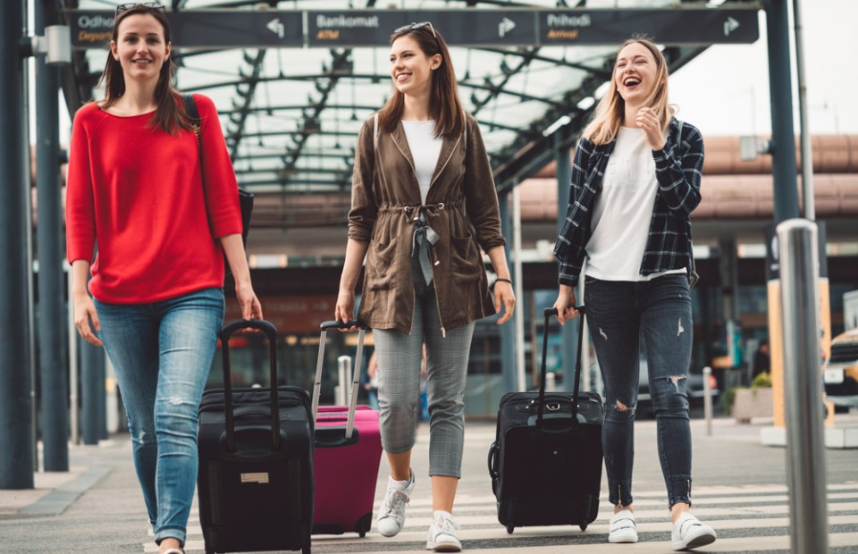 Drei junge Frauen in Freizeitkleidung gehen lachend mit ihren Koffern über einen Zebrastreifen vor einem Bahnhofsgebäude.