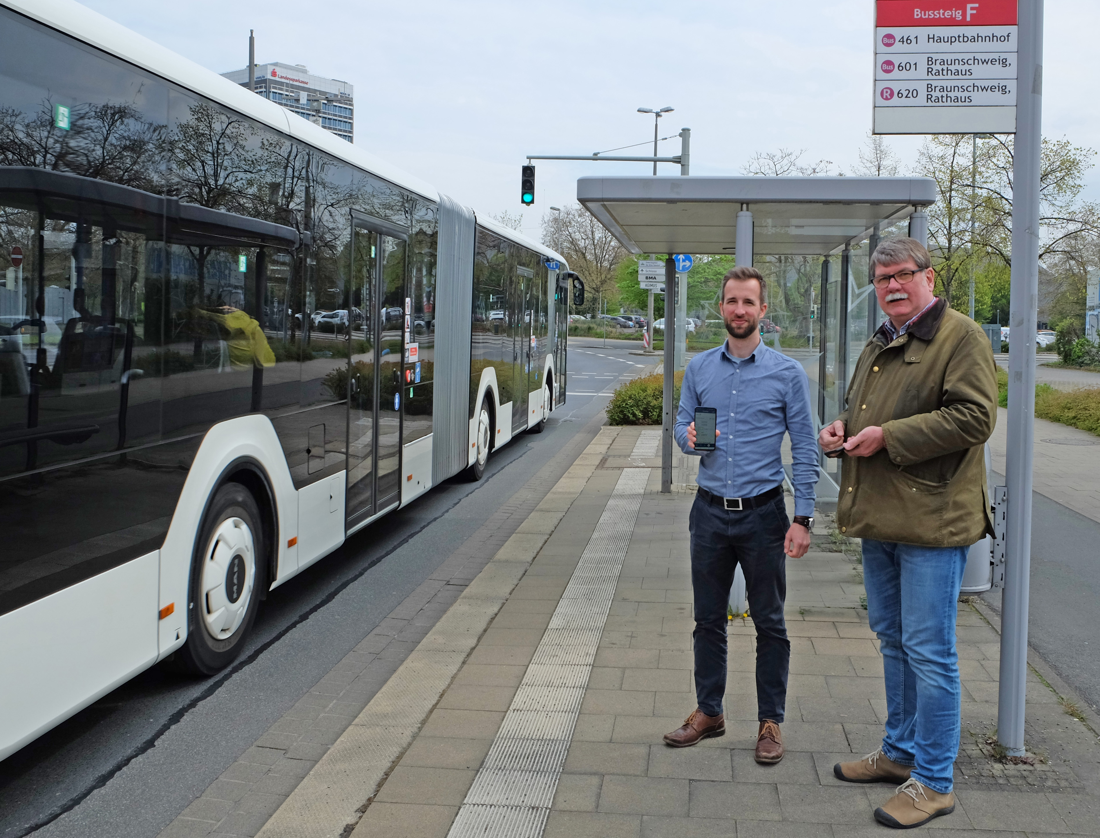 Zwei Männer stehen an einer Bushaltestelle. Einer der Männer hält sein Smartphone in Richtung Kamera.. Auf dem Smartphone ist das Deutschlandticket in der VRB-App zu sehen.